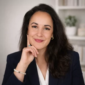 Avocate aux cheveux foncés ondulés, tête appuyée sur la main, léger sourire, vêtue d’un blazer bleu, dans un environnement intérieur clair.
