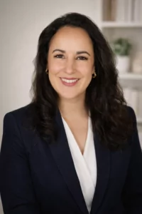Avocate souriante aux cheveux foncés ondulés, portant un blazer bleu marine et une blouse blanche, dans un portrait professionnel sur fond neutre.